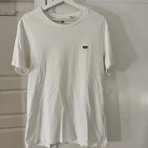 Vit t-shirt från Levi's i bomull - Klassisk vit t-shirt från Levi's med liten röd logotyp på bröstet. Fläckar i armhålan som syns lite men inget märkbart. T-shirten har rund halsringning och korta ärmar. Tillverkad i mjuk bomull för en skön känsla. Perfekt basic-plagg med clean look.