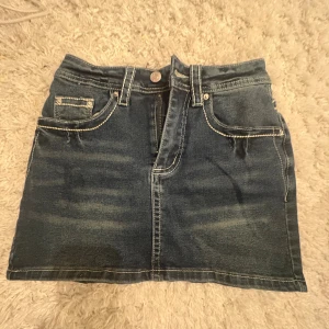 Mörkblå jeanskjol med broderi från Romwe - Säljer en mörkblå jeanskjol från Romwe i storlek XS. Kjolen har klassisk femficksdesign, kontrastsömmar och snygga broderade fjärilar med pärldetaljer på bakfickorna. Modellen är kort och har dragkedja framtill. 