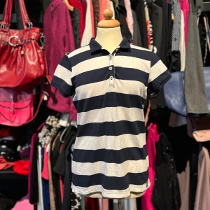 Randig pikétröja från HV Society - Snygg pikétröja från HV Society i marinblått och vitt med breda horisontella ränder. Klassisk krage, tre knappar framtill och korta ärmar. Broderad logga i nacken och små slitsar i sidan för lite extra stilpoäng.