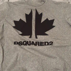 Grå Dsquared2 t-shirt med svart tryck - Snygg grå t-shirt från Dsquared2 med stort svart lönnblads-tryck och logga på bröstet. Klassisk rund hals och korta ärmar. Perfekt för dig som gillar streetwear och vill sticka ut med ett coolt designerplagg.