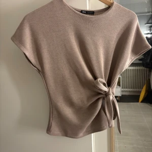 Beige topp med knyt från Zara - Säljer en beige topp från Zara med korta ärmar och rund hals. Toppen har en snygg knytning i sidan som ger en cool och trendig look. Materialet är mjukt och lätt ribbat, perfekt för en stilren outfit.