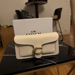 Beige kuvertväska från Coach - Stilren beige kuvertväska från Coach med exklusiv känsla. Väskan har en stor guldfärgad C-detalj på framsidan och är tillverkad i strukturerat skinn. Perfekt storlek för det viktigaste och levereras med originalbox.