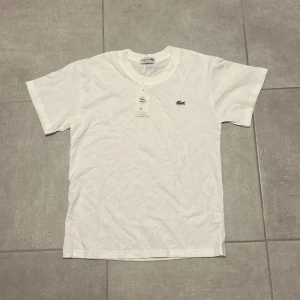 Vit t-shirt från Lacoste - Klassisk vit t-shirt från Lacoste med rund halsringning och den ikoniska krokodilloggan broderad på bröstet. Tillverkad i mjuk bomull och har en normal passform. Perfekt basic-plagg för en clean och stilren look.