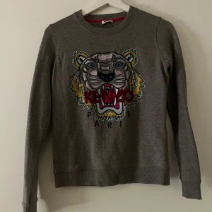 Grå Kenzo sweatshirt med tiger - Säljer en grå sweatshirt från Kenzo med ikoniskt broderat tigerhuvud och röd Kenzo-logga på bröstet. Tröjan har rund hals, långa ärmar och ribbade muddar. Perfekt statement piece för dig som gillar streetstyle. Stl XS