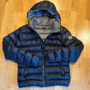 Mörkblå Colmar dunjacka med huva - Snygg mörkblå dunjacka från Colmar med quiltad design och huva. Jackan har dragkedja framtill, två sidofickor med dragkedja och en rund Colmar-logga på ärmen. Insidan är grå och materialet är lätt och värmande, perfekt för kyliga dagar. Jackan är i väldigt fint skick, använd 2 gånger. Skriv om du har frågor. 