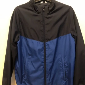 Svart och blå vindjacka stl 164 - Snygg tvåfärgad vindjacka i svart och blått från C. Jackan har dragkedja framtill, ståkrage och två fickor. Perfekt för blåsiga dagar och enkel att matcha med det mesta. Lätt och smidig modell som funkar året runt.