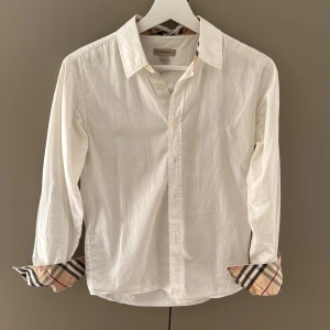 Vit Burberry skjorta med rutiga detaljer - Vit skjorta från Burberry med logga på bröstet. Skjortan är i storlek 164 vilket motsvarar Xs, liten missfärgning på insidan av ena ärmen dock inget man tänker på.