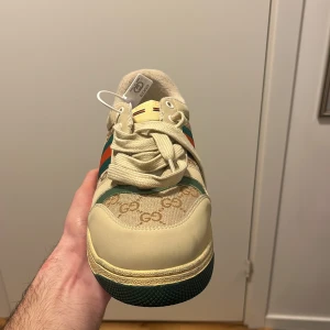 Gucci sneakers med GG-mönster - Snygga sneakers från Gucci i beige med klassiskt GG-mönster på sidan och gröna samt röda ränder. Skon har snörning, rund tå och detaljer i grönt på hälen med guldfärgad Gucci-logga. Materialmix av skinn och textil, tillverkad i Italien.