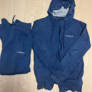 Marinblå set från McKenzie - Säljer en marinblå hoodie från McKenzie med dragkedja och snörning i huvan. Tröjan har ribbade muddar och en liten logga på bröstet. Materialet är mjuk bomull och polyester, perfekt för chill dagar. Huvan är fodrad i grått för extra stilpoäng. Sedan kommer den med byxor i samma färg och material(byxorna är i storlek M, alltså en storlek större än koftan då modellen på byxorna är mindre än vanligt)