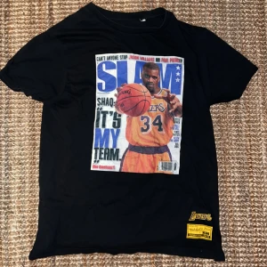 Slam Shaq  t-shirt Mitchell & Ness - Svart t-shirt från Mitchell & Ness med stor färgglad print av Shaquille O'Neal i Lakers-tröja på framsidan, inspirerad av SLAM Magazine. Lakers-logga och gul etikett nertill. 100% bomull, klassisk passform och korta ärmar. Perfekt för basketfans! Passar M och S