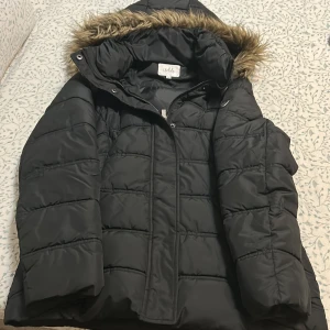 Svart pufferjacka med fuskpäls från Isolde - Svart pufferjacka från Isolde i storlek 38. Jackan har huva med fuskpäls, dragkedja och tryckknappar framtill samt två sidofickor. Quiltad design och långärmad modell, perfekt för kalla dagar. Materialet är syntet och polyester.