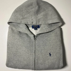 Polo Ralph Lauren Grå Zip Hoodie - Detta är en Polo Ralph Lauren hoodie i mjuk grå färg. Den har dragkedja fram och den kända Polo-loggan på bröstet. Tröjan är gjord av mjukt och skönt tyg som är bekvämt att ha på sig varje dag.