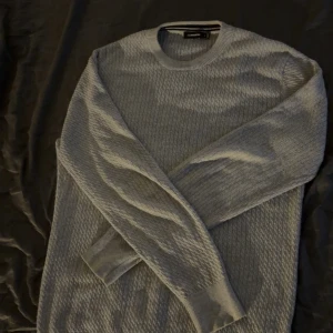 Grå stickad tröja från J.Lindeberg - Snygg grå stickad tröja från J.Lindeberg med diskret mönster och rund halsringning. Tröjan har ribbade muddar vid ärmslut och nederkant, samt en liten logotagg i sidan. Perfekt för lager-på-lager under kyliga dagar.