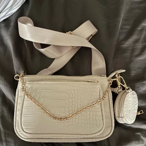 Beige axelväska med kedja och croco-mönster - Snygg beige axelväska med croco-mönstrad yta, guldfärgade detaljer och kedja framtill. Väskan har flera fack med dragkedja och en bred justerbar axelrem. Dessutom medföljer en liten matchande miniväska som sitter fast i sidan.