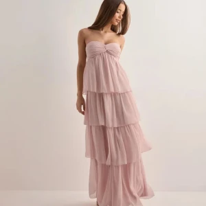 Ljusrosa långklänning med volanger - Säljer en romantisk ljusrosa långklänning med flera lager volanger och knytdetalj vid bysten. Klänningen är axelbandslös men kan stylas med halterneck, och har en flowy siluett i tunt, luftigt material. Perfekt för sommarens alla tillfällen.