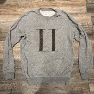 Grå sweatshirt från Les Deux - Säljer en stilren grå sweatshirt från Les Deux med stort II-tryck på bröstet. Tröjan har rund hals, ribbade muddar och är tillverkad i mjuk bomull. Perfekt för en clean och avslappnad look.