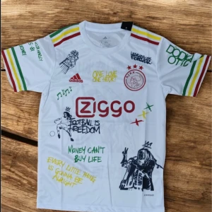Ajax-Bob Marley vit fotbollströja  - Vit Ajax fotbollströja från Adidas i slim fit med Bob Marley-tryck och graffitiinspirerade citat. Färgglada ränder i rött, gult och grönt på ärmarna, klubbmärke och Ziggo-logga framtill. Materialet är lätt och andas, perfekt för match eller träning.