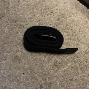 Svart flätat bälte med skinn-detaljer - Snyggt svart flätat bälte med klassiskt spänne i metall och detaljer i svart skinn vid ändarna. Bältet har en stilren look och passar perfekt till jeans eller chinos. Flätad design gör det enkelt att justera längden efter behov.