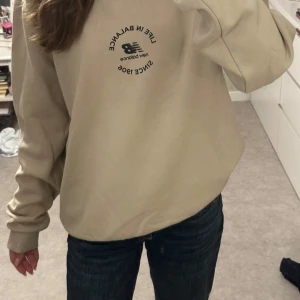Beige sweatshirt från New Balance - Säljer en beige sweatshirt från New Balance med svart tryck på bröstet där det står 'Life in Balance Since 1906'. Tröjan har rund hals, långa ärmar och ribbade muddar. Perfekt för en chill och avslappnad stil.