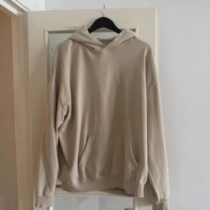Beige hoodie med tryck "Tröjan du hatar" - Beige hoodie i storlek L med stor vit text "Tröjan du hatar" på ryggen. Klassisk känguruficka framtill, huva och avslappnad passform. Perfekt för dig som gillar streetstyle och vill sticka ut med ett unikt statement-plagg.