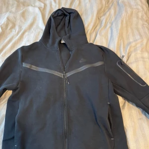 Svart Nike hoodie med dragkedja - Svart hoodie från Nike med hel dragkedja och huva. Snygga detaljer med gråa ränder över bröstet och en extra ficka med dragkedja på ärmen. Perfekt för chill eller träning, klassisk och stilren design.