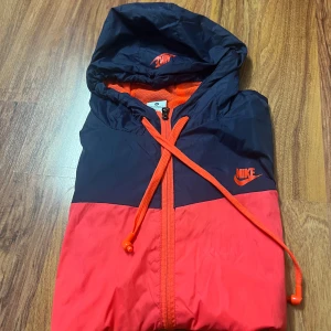Nike vindjacka i röd och marinblå - Nike vindjacka med huva i röd och marinblå färg. Jackan har dragkedja framtill, två fickor med dragkedja och justerbar dragsko i huvan. Lättviktig och sportig design med meshfoder på insidan och klassisk Nike-logga på bröstet.