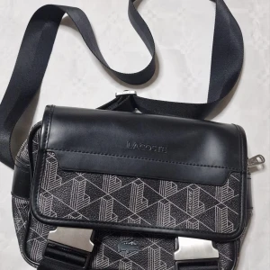 Svart axelväska från Lacoste - Snygg svart axelväska från Lacoste med geometriskt mönster och silverfärgade detaljer. Väskan har justerbar rem, lock med präglad logga och smarta fack. Materialmix av skinn och syntet ger en modern vibe.