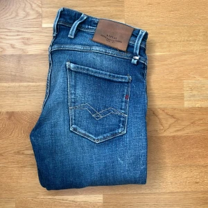 Replay Anbass slim fit jeans blå - Snygga blå Replay Anbass jeans i slim fit med klassisk femficksdesign och coola slitningar. Jeansen har medelblå tvätt, kontrastsömmar och läderpatch bak i midjan. Perfekta för digsom diggar en stilren look.