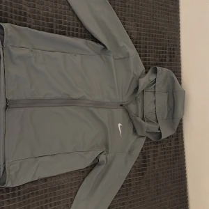 Grå vindjacka från Nike med luva  - Snygg grå vindjacka från Nike med huva och hel dragkedja framtill. Jackan har lång ärm med resår vid ärmslut och en stilren vit Nike-logga på bröstet. Perfekt för blåsiga dagar och sportiga outfits. Priset kan alltid diskuteras!!