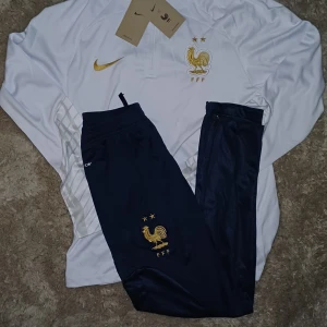 Frankrike Nike Set - Säljer en Frankrike Set-Nike i vitt med guldiga detaljer och broderad tupp-logga. Tröjan har halv dragkedja och diskret rutmönster på sidorna. Materialet är lätt och andas, perfekt för träning eller match.