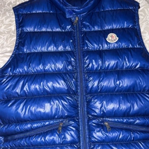 Blå dunväst från Moncler - Snygg blå dunväst från Moncler med glansig finish och klassisk logga på bröstet. Västen har två dragkedjefickor framtill och hög krage. Perfekt för lager-på-lager och ger en sportig vibe till din outfit. Bud