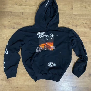 Svart hoodie med tryck från H&M - Svart hoodie från H&M med relaxed fit och coolt tryck på bröstet i orange och vitt. Snygga vita grafiska texter längs ärmarna och huva med snörning. Perfekt för dig som gillar streetstyle och vill sticka ut lite extra.