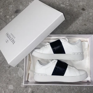 Valentino Rockstud vita sneakers - Snygga vita Valentino Rockstud sneakers med svart band och ikoniska nitar över vristen. Skorna är i skinn med rund tå, platt sula och vita snören. Använd 2 gånger och kommer i originalkartong.