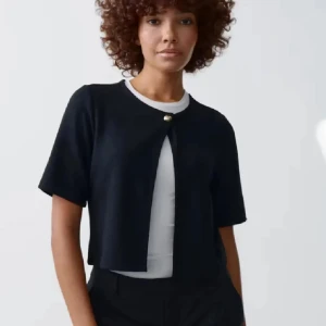 Svart kort kofta med knapp - Kort svart kofta med korta ärmar och en snygg guldfärgad knapp framtill. Koftan har en rak passform och är perfekt att slänga över en t-shirt. Enkel och stilren design som passar till många outfits.