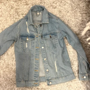 Ljusblå jeansjacka med slitningar - Säljer en ljusblå jeansjacka från Gina Tricot med coola slitningar och klassiska bröstfickor med knappar. Jackan har en rak passform, silverfärgade knappar och långa ärmar. Perfekt för dig som gillar en trendig stil.