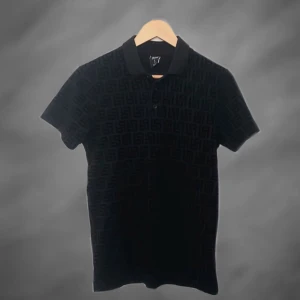 Svart mönstrad pikétröja från Siksilk - Svart pikétröja från Siksilk med diskret mönster över hela tyget. Klassisk krage, knäppning med tre knappar och korta ärmar. Snygg och stilren design som passar perfekt till jeans eller shorts.