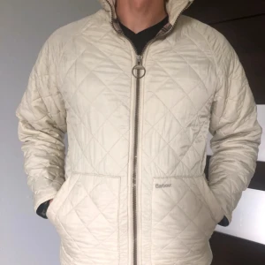 Beige quiltad jacka från Barbour - Quiltad beige jacka från Barbour med hög krage, två stora fickor framtill. Jackan har ett klassiskt rutmönster och är perfekt för dig som gillar stilren och tidlös design. Perfekt inför hösten!