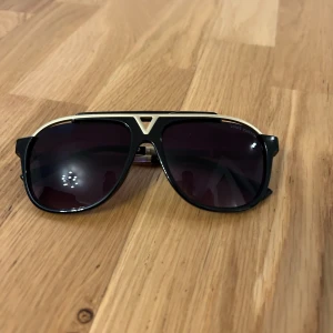 Louis Vuitton svarta och guldiga solglasögon - Snygga solglasögon från Louis Vuitton med svart och guldig båge i klassisk aviator-stil. Ljuslila tonade glas och diskret LV-logga på skalmarna. Metall- och plastdetaljer ger en lyxig känsla och unik design.