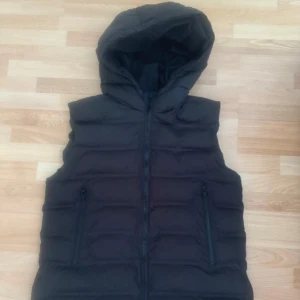 Svart dunväst med huva från Zara - Svart dunväst från Zara med huva och två fickor med dragkedja. Västen har quiltad design och stängs med en kraftig dragkedja framtill. Perfekt för lager-på-lager och chill stil. Passar dig som gillar enkel och clean look. Det är storlek 164cm, men kan passa xs/s också. Använd en gång. Bröst 104cm.