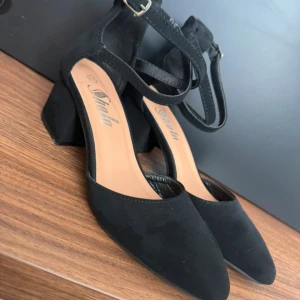 Svarta pumps med klack från Shela - Stilrena svarta pumps från Shela med spetsig tå och bred klack. Skorna har en snygg rem med spänne runt ankeln och är tillverkade i mockaliknande material. Perfekta för dig som vill ha en klassisk och trendig look.