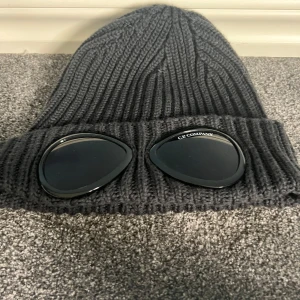Svart C.P. Company Goggle mössa - Svart ribbstickad mössa från C.P. Company med ikoniska goggle-linser på uppviket. Tillverkad i 100% ull för extra värme och komfort. Mössan har en klassisk form med hög uppvikning och tydlig logga på ena linsen. Perfekt för dig som vill sticka ut med streetwear-vibe.
