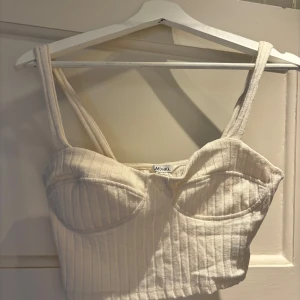 Ribbad vit korsettopp från Monki - Superfin ribbad korsettopp från Monki i offwhite. Toppen har markerade kupor, breda axelband och croppad passform. Perfekt att styla med jeans eller kjol för en trendig look. Materialet känns mjukt och stretchigt.