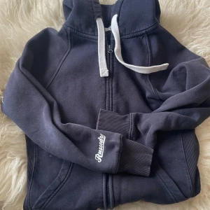 Mörkblå hoodie från Russedress S - Mörkblå hoodie från Russedress med vit snörning och dragkedja framtill. Tröjan har huva, ribbade muddar och tryckt logga på ärmen. Perfekt för kalla dagar och chill vibes.
