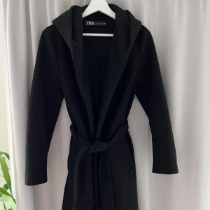 Svart kappa med huva från Zara - Varm kappa från Zara med huva och knytskärp i midjan. Funkar att ha på vintern. Lång modell med rak passform. Använd ett par gånger i vintras. Säljer då den inte är min stil längre. Bilderna är lånade, min har inga noppor alls. Så typ nytt skick.