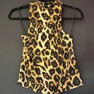 Leopardmönstrad ärmlös linnetopp - Snygg ärmlös linnetopp med leopardmönster i svart, beige och bruna toner. Toppen har hög hals och smala axelband, perfekt för dig som vill sticka ut med ett djurmönster. Materialet känns mjukt och följsamt mot huden.