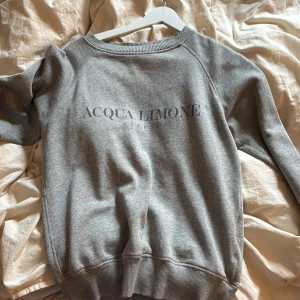 Grå sweatshirt Acqua Limone XS - Grå sweatshirt från Acqua Limone i storlek XS. Klassisk modell med rund hals, ribbade muddar och stort gult citrontryck på ryggen samt logga framtill. Perfekt för chill dagar och enkel att matcha med jeans eller leggings.