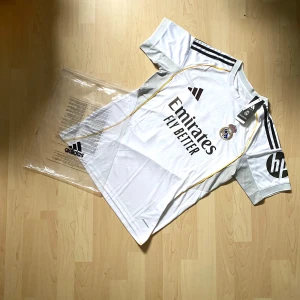 Real Madrid Mbappé 10 fotbollströja Adidas - Real Madrid fotbollströja med Mbappé och nummer 10 på ryggen. Vit med svarta detaljer, gula linjer och klubbmärke på bröstet. Adidas-logga, HP-logga på ärmen och 'Emirates Fly Better' tryck på framsidan. Tillverkad i lätt funktionsmaterial, kortärmad modell.