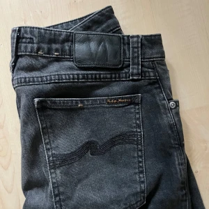 Svarta jeans från Nudie Jeans - Säljer ett par svarta jeans från Nudie Jeans i storlek W31 L32. Jeansen har klassisk femficksdesign, svarta sömmar på bakfickorna och Nudie Jeans-logga på bakfickan och knappen. Materialet är jeans i bomull och passformen är tight och modern.