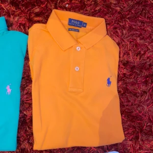 Orange polo t-shirt från Ralph Lauren - Snygg orange polo t-shirt från Ralph Lauren i modellen The Skinny Polo. Klassisk krage, två knappar och blå broderad logga på bröstet. Perfekt för dig som gillar färg och stilren design. Gjord i mjuk bomull som känns skön mot huden.