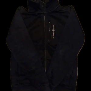 Svart hoodie från Sail Racing med dragkedja - Svart hoodie från Sail Racing med hel dragkedja framtill och broderad logga på bröstet. Tröjan har huva, lång ärm och är tillverkad i mjukt fleecematerial. Perfekt för kyliga dagar och enkel att matcha med andra plagg.